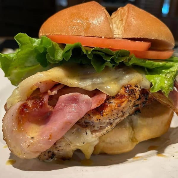 A grilled chicken sandwich with melted cheese, ham, lettuce, and tomato on a bun.