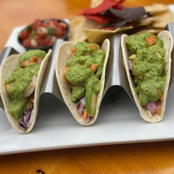 Three shrimp tacos with green sauce, avocado, and red onion, served with salsa and chips.