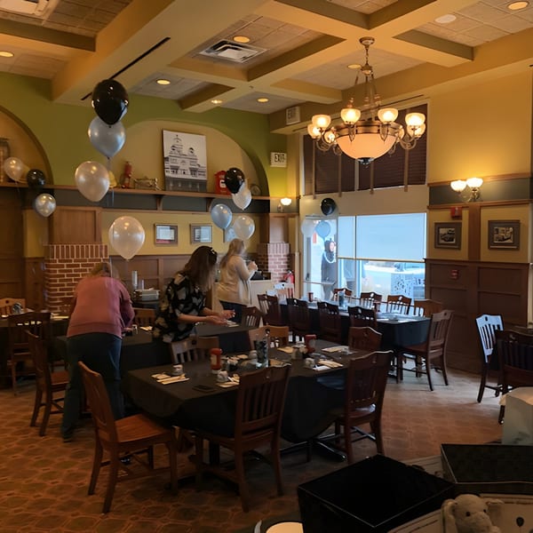 Restaurant dining room with black and white balloons, people setting tables for an event.