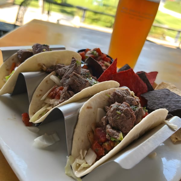Three steak tacos with pico de gallo and onions, served with chips and a beer.