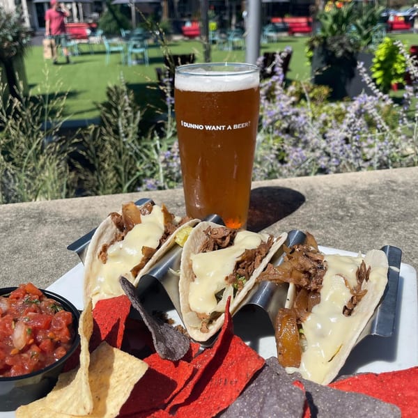 Three tacos with melted cheese and meat, chips, salsa, and a beer on an outdoor patio.