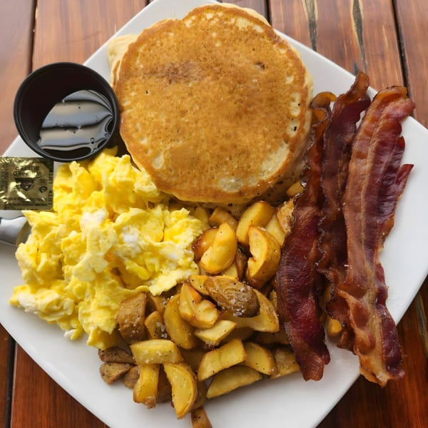 A breakfast platter with pancakes, scrambled eggs, home fries, bacon, and a side of syrup.