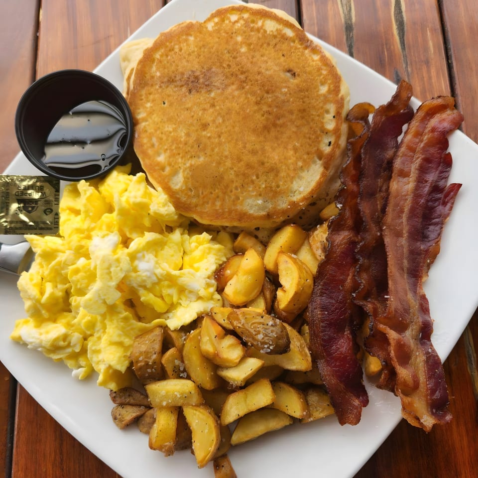 A breakfast platter with pancakes, scrambled eggs, home fries, bacon, and a side of syrup.